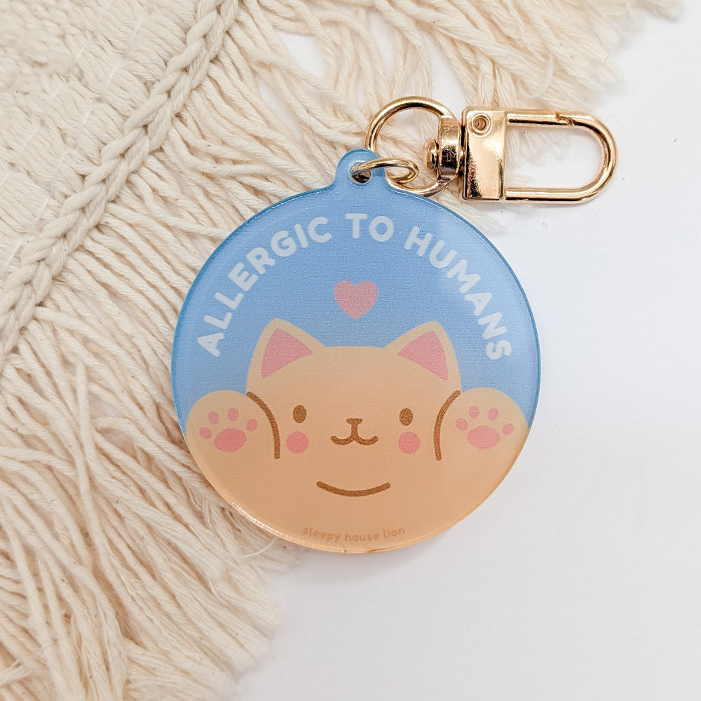 A round keychain tag with a light blue top half and a peach-colored cat face on the bottom half, attached to a gold clasp. The tag reads 'ALLERGIC TO HUMANS' with a small pink heart above the cat's head.