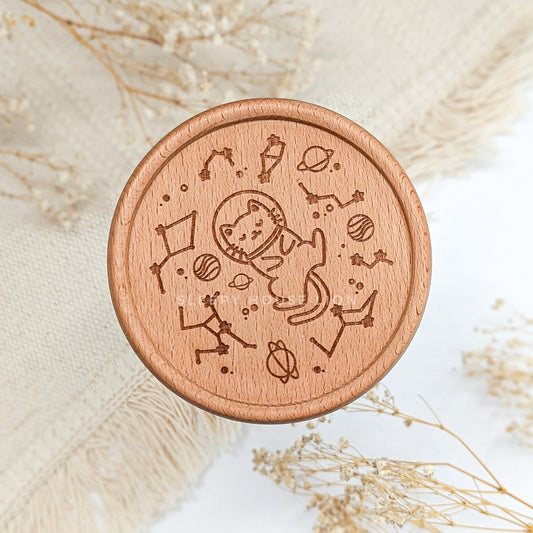Wooden coaster with engraved design of a cat astronaut on a textured surface with dried plants.