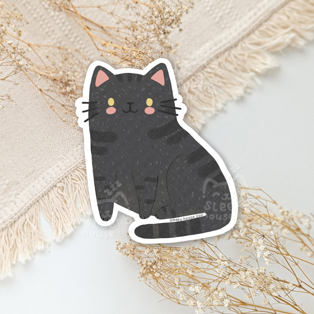 Sticker of a black cat with pink ears on a textured surface with dried flowers.