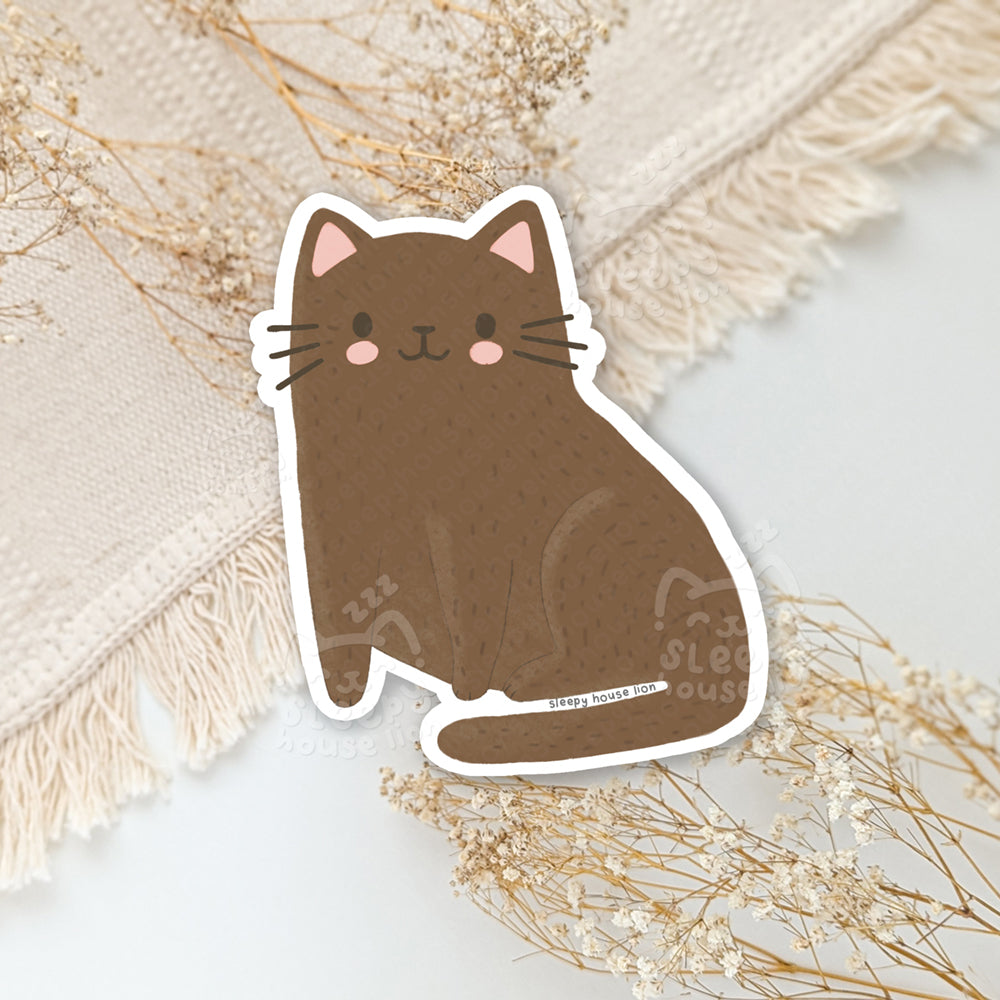 Sticker of a brown cat with pink accents on a light background with dried flowers.