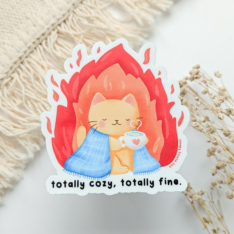 cozy cat under blanket with flames around with text: totally cozy, totally fine.