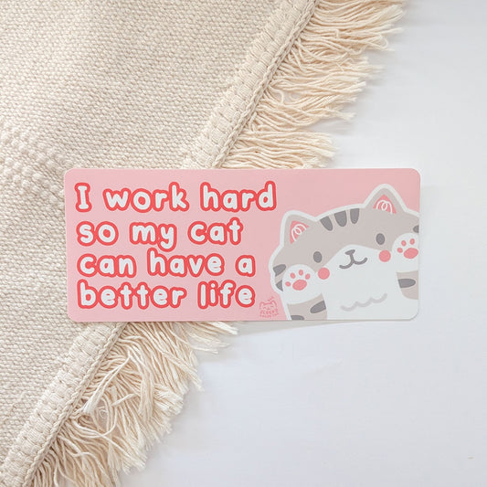 Pink sticker with cat illustration and text "I work hard so my cat can have a better life" on a textured surface