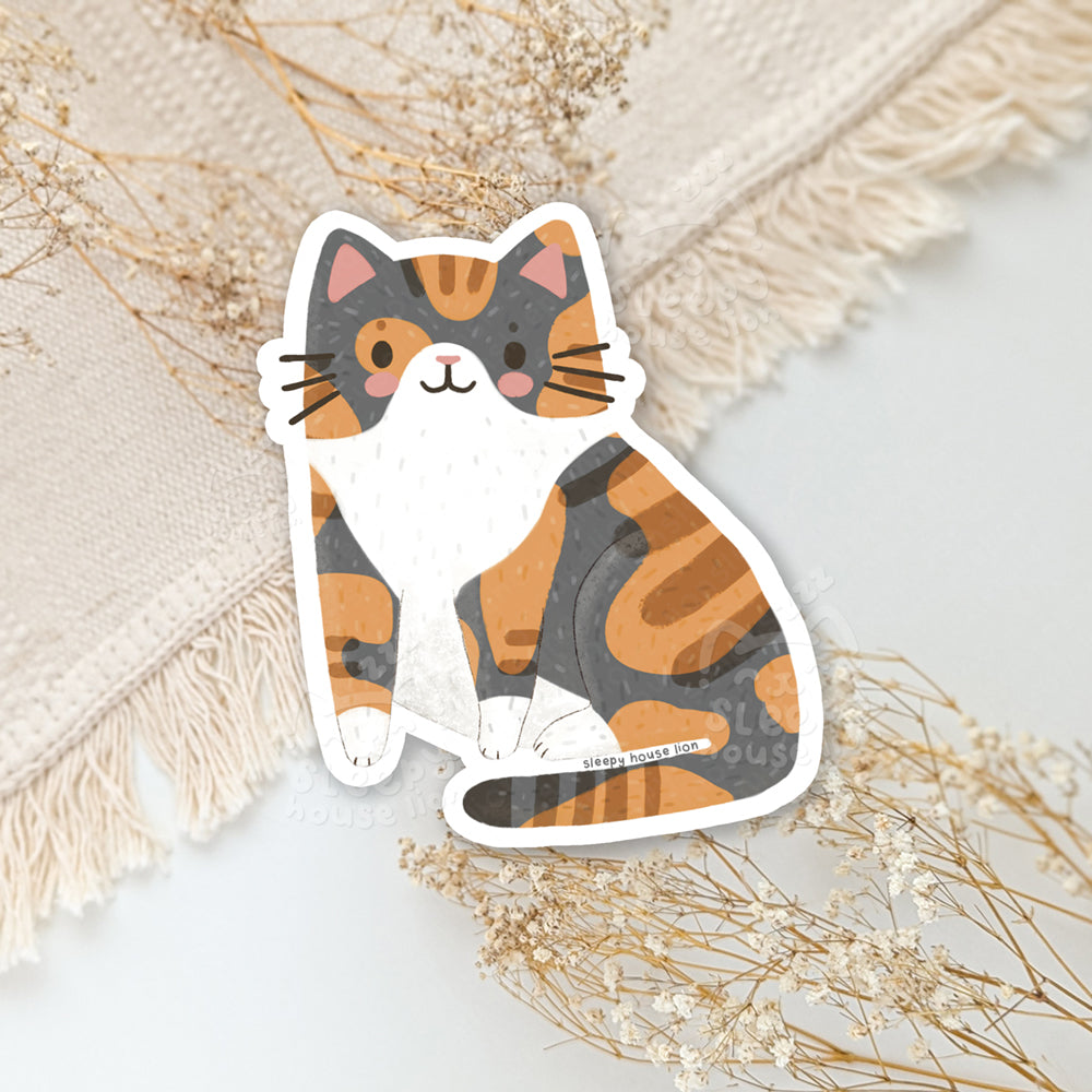 Calico cat sticker on a textured surface with dried flowers