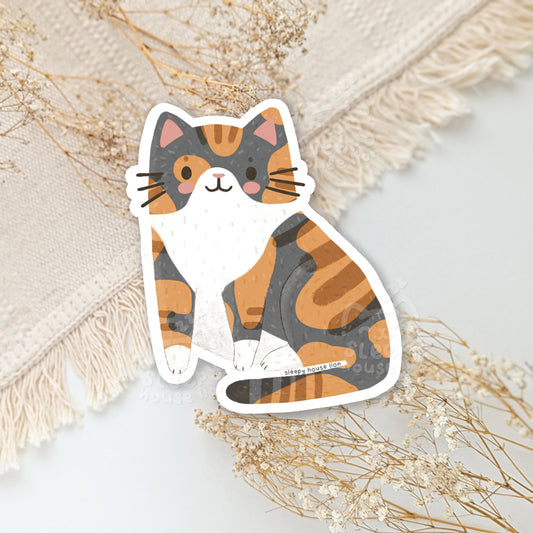 Calico cat sticker on a textured surface with dried flowers