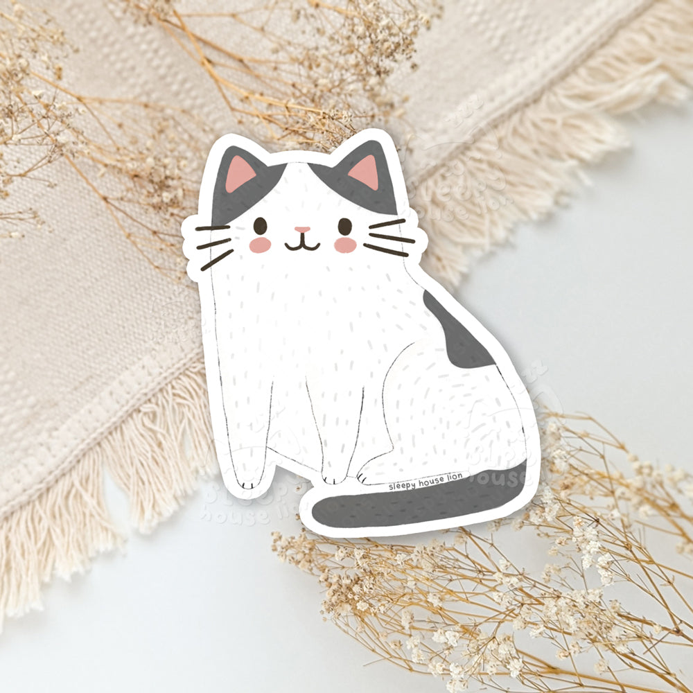 Sticker of a white cat with gray ears and tail on a textured surface with dried flowers.