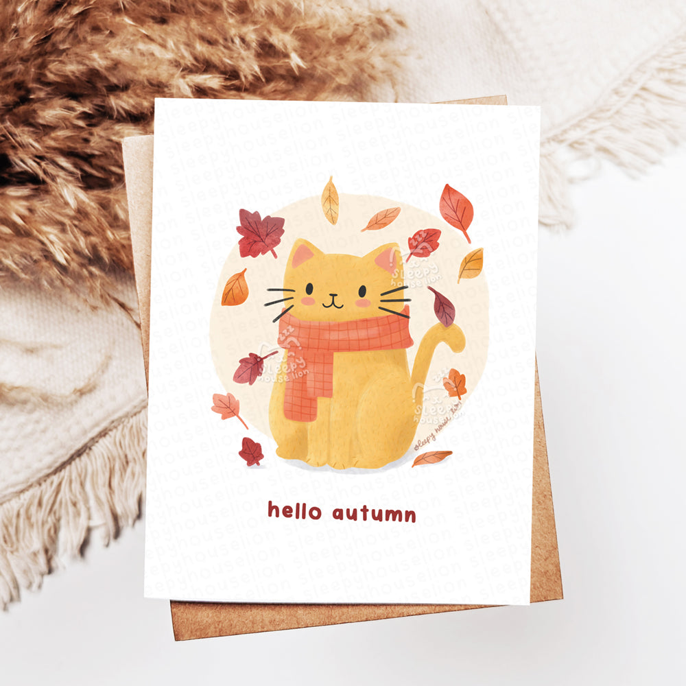 Card with a cat illustration wearing a scarf and autumn leaves, on a textured surface.