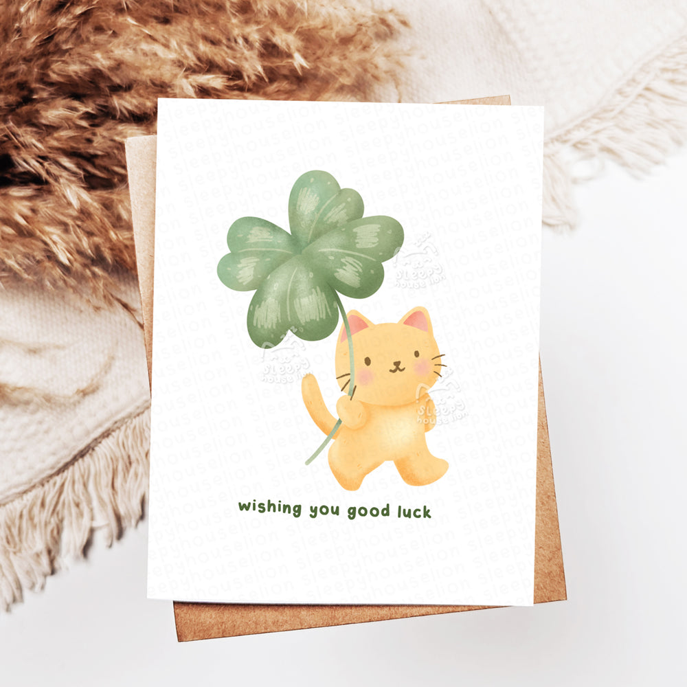 Greeting card with a cat holding a four-leaf clover and text 'Wishing you good luck' on a textured surface.